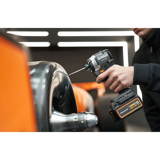 Person uses a DEWALT power tool to work on a car part in an indoor setting with orange and black details.