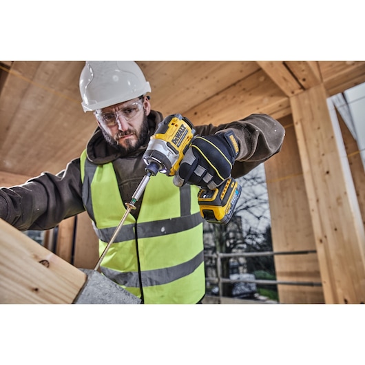 A person wearing a hard hat, high-visibility vest, and protective gloves uses a DEWALT cordless impact driver to fasten a screw into a wooden beam at a construction site.