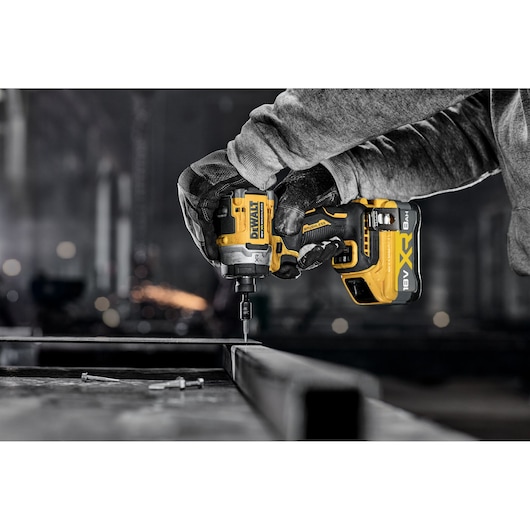 A person wearing gloves is using a yellow and black DEWALT 18V XR cordless impact driver to drill or fasten a screw into a metal frame in an industrial workshop setting.