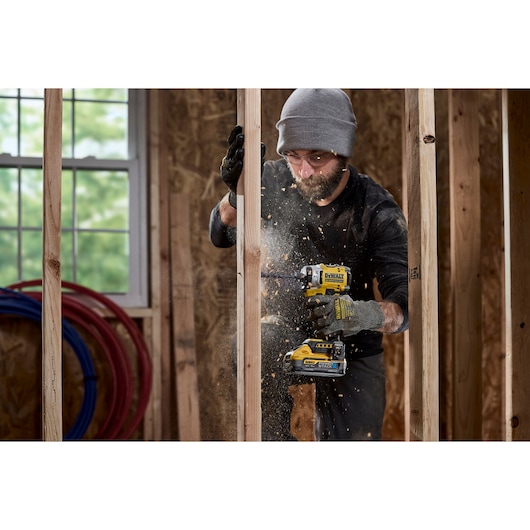 A person wearing gloves and a beanie uses a DEWALT DCF860N cordless impact driver on a wooden frame while sawdust is flying. The setting appears to be a construction site with wooden beams and a window in the background.