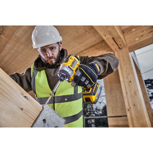 A construction worker wearing a safety helmet and high-visibility vest is using a DEWALT DCF860N cordless impact driver to drive a screw into a wooden structure. The worker is also wearing gloves. The setting appears to be an unfinished building with wooden beams.