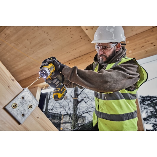 A person wearing a safety helmet and high-visibility vest uses a DEWALT cordless impact driver to drill a screw into a metal bracket attached to a wooden beam at a construction site.