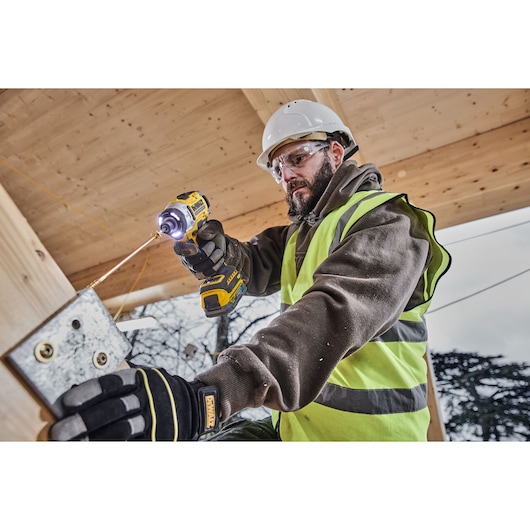 A person wearing a safety helmet and high-visibility vest uses a DEWALT DCF860N power tool to drive a screw into a metal plate on a wooden structure at a construction site.