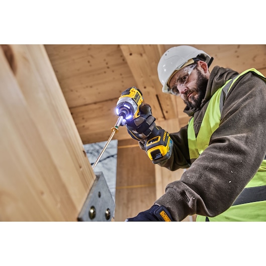 A person wearing a safety helmet and high-visibility vest is using a DEWALT DCF860N cordless impact driver to fasten a screw into a large wooden beam, in a construction setting.