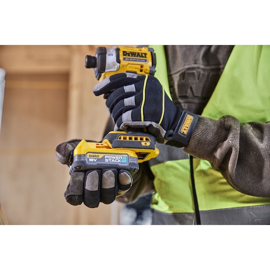 A person wearing work gloves and a high-visibility vest holding a DEWALT 18V POWERSTACK cordless impact driver in a workshop environment.