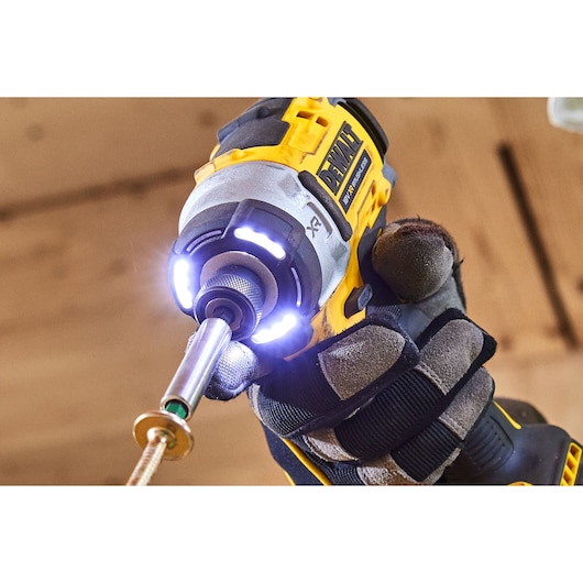 A close-up of a person wearing work gloves using a DEWALT DCF860N cordless impact driver with built-in LED lights, driving a screw in a wooden surface.