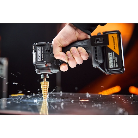 Hand uses DEWALT cordless drill with step bit to drill into metal, metal shavings scattered on surface.