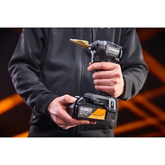Person holding DEWALT cordless drill and battery pack, McLaren edition, in a workshop setting.