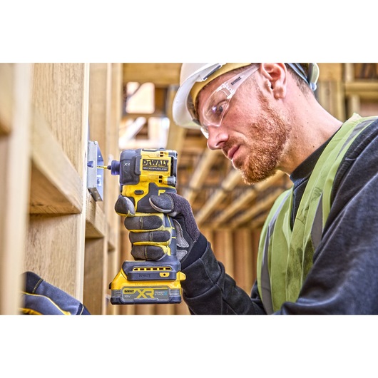 Worker uses DEWALT DCF870E2T impact driver on a wood frame, wearing gloves, helmet, and safety vest.