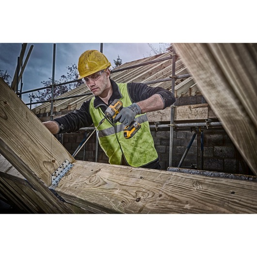 A construction worker wearing a yellow hard hat and reflective vest uses a DEWALT cordless drill to fasten metal brackets on wooden beams at a building site. His face is blurred.