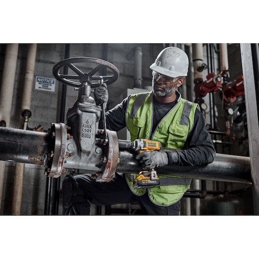 A worker wearing a safety vest, gloves, and helmet is using a yellow DEWALT DCF891H2T impact wrench on a large industrial valve. The scene appears to be inside a facility with metal pipes and machinery.