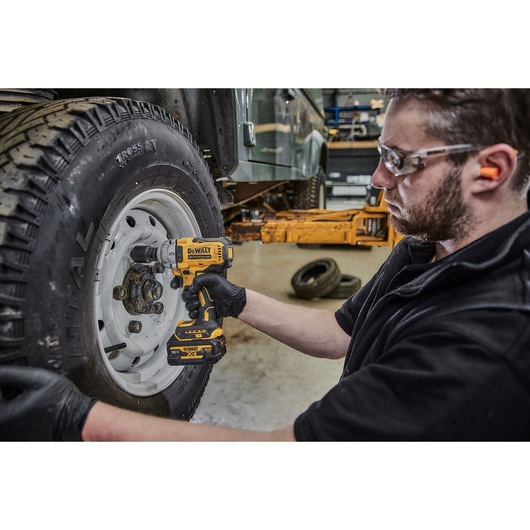 A person wearing gloves uses a DEWALT DCF891NT cordless impact wrench to work on the wheel of a vehicle in a garage. The