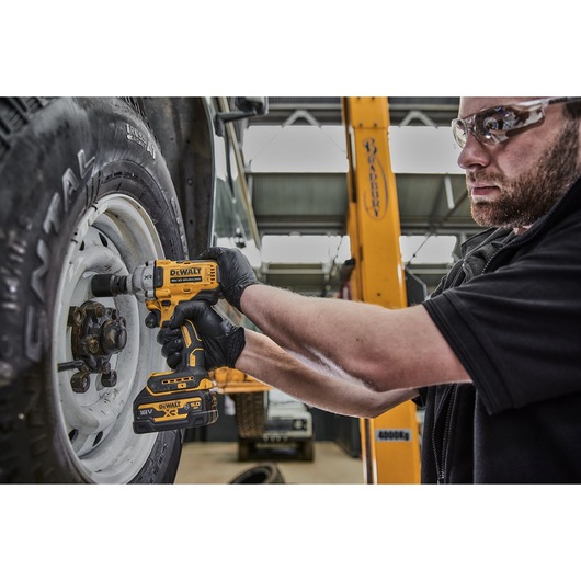 A person wearing black gloves uses a DEWALT 18V cordless impact wrench (model DCF891N) to remove a lug nut from a vehicle's