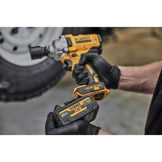 A person wearing black gloves is holding a yellow and black DEWALT cordless impact wrench, model DCF891N, and an 18V XR