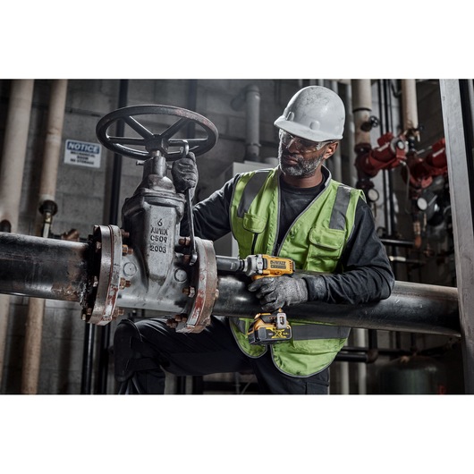 A worker wearing a safety helmet and green high-visibility vest is using a DEWALT DCF891N cordless impact wrench to tighten