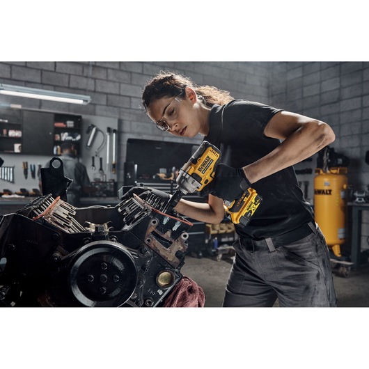 A person using a DEWALT cordless impact wrench (model DCF891N) to work on an engine in a workshop environment. The person is