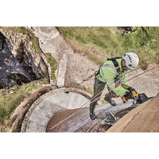 A worker wearing safety gear and a helmet is secured with ropes while using a DEWALT power tool to work on the side of a tall structure outdoors. The scene is taken from above, showing the worker high above the ground.