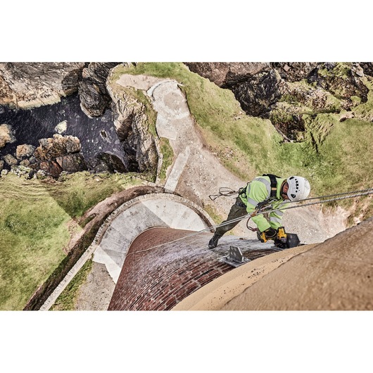 A person wearing safety gear is using a DEWALT power tool while secured with ropes, working on the exterior of a tall brick tower or structure, with a rocky landscape and water visible below.