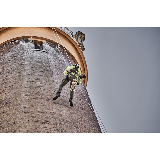 A worker in safety gear is suspended by ropes, performing maintenance or inspection on the exterior of a tall brick tower.