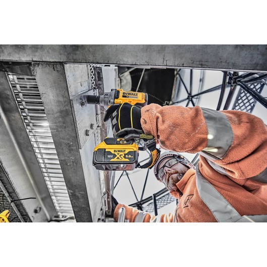 A worker in safety gear uses a DEWALT 18V XR cordless impact wrench (model DCF894P2) to fasten bolts on a metal structure at a construction site.
