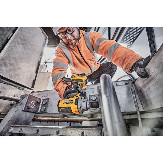 A construction worker wearing an orange high-visibility jacket and gloves is using a DEWALT DCF894P2 cordless impact wrench to tighten bolts on a metal structure at an industrial site.