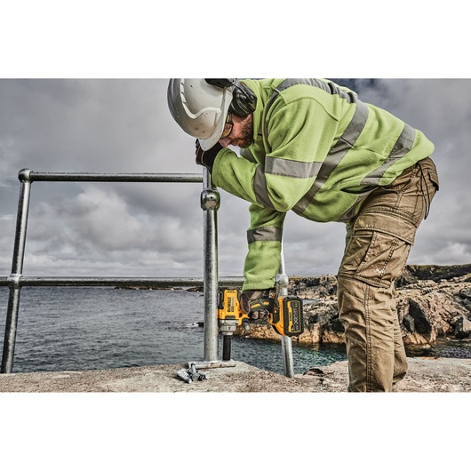 A worker wearing safety gear, including a helmet and high-visibility jacket, uses a DEWALT cordless impact wrench to fasten bolts on a metal railing outdoors near a body of water.