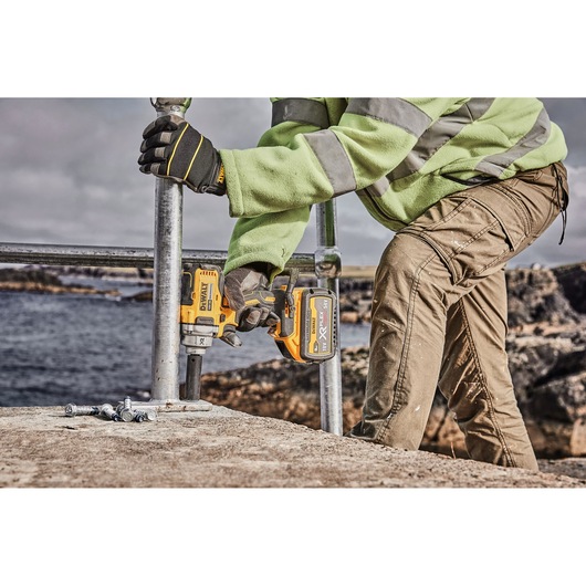 A person wearing work gloves and a high-visibility jacket is using a DEWALT DCF894P2 cordless impact wrench to secure metal pipes outdoors, near a body of water.