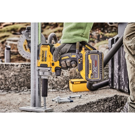 A DEWALT cordless impact wrench (model DCF894P2) being used to fasten a bolt to a metal base outdoors. The tool is held by a person wearing work gloves and a green jacket. Other construction tools and bolts are visible in the background.