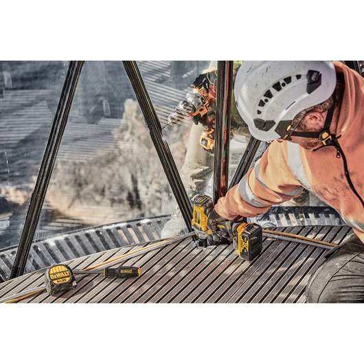 A construction worker wearing a safety helmet and high-visibility jacket uses a DEWALT cordless impact wrench (model DCF894P2) to fasten bolts on a metal structure. Other DEWALT tools and accessories are placed on the ground nearby.