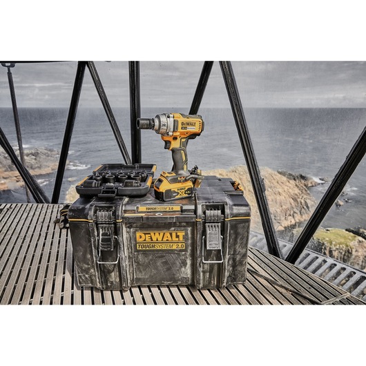 A DEWALT cordless impact wrench and battery are placed on top of a DEWALT ToughSystem 2.0 tool storage box, with drill bit accessories, set outdoors near a window overlooking a rocky coastline.