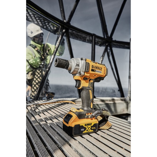 A DEWALT DCF894P2 cordless impact wrench with an 18V XR Li-Ion 5.0Ah battery, placed on a wooden slatted surface, with an industrial structure and a person in safety gear in the background.