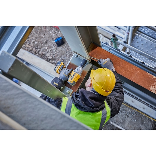A person wearing a yellow hard hat and high-visibility vest uses a DEWALT cordless impact wrench to fasten bolts on a steel beam at a construction site.