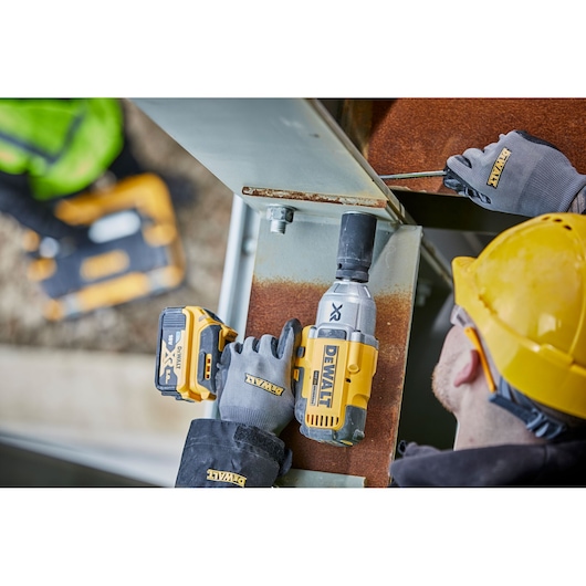 A worker wearing a yellow hard hat and DEWALT gloves uses a DEWALT cordless impact wrench (model DCF897N) on a steel beam at a construction site.