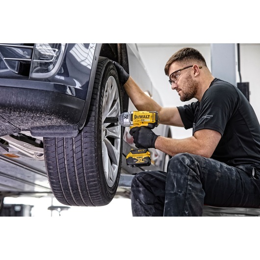 A person with a blurred face is using a DEWALT DCF899N cordless impact wrench to work on the wheel of a car, wearing black
