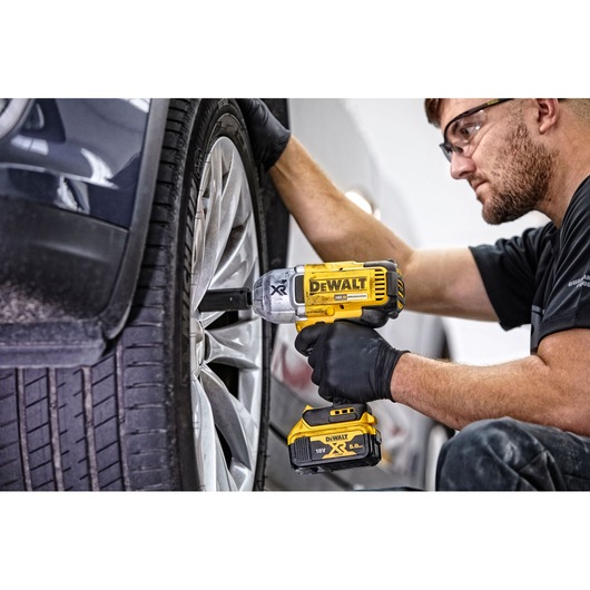 A person using a DEWALT DCF899N cordless impact wrench to loosen or tighten the lug nuts on a car wheel. The tool is yellow