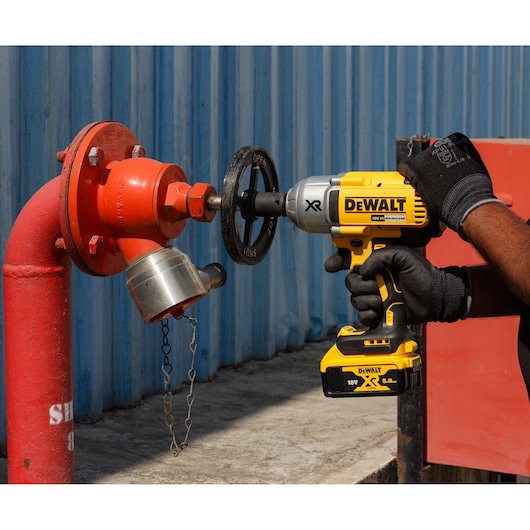 A person wearing gloves is using a DEWALT cordless impact wrench (model DCF899P2-QW) to adjust a valve on a red industrial pipe near a blue metal wall.