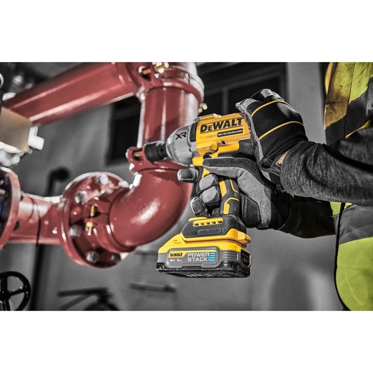 A gloved hand uses a yellow DEWALT cordless impact wrench near large red industrial pipes indoors.