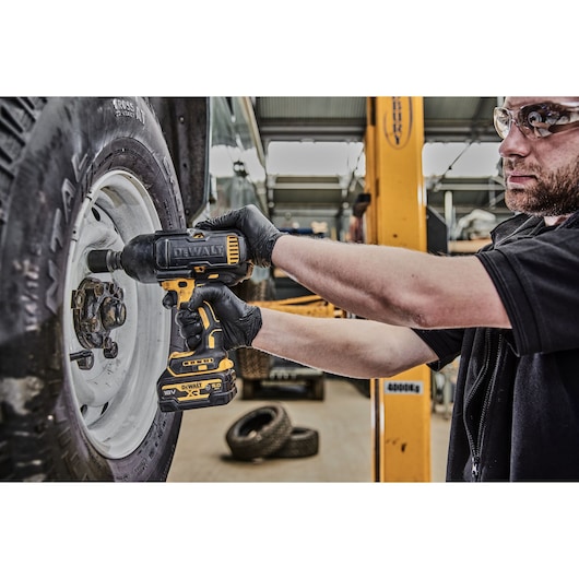 A person wearing black gloves using a DEWALT DCF900P2G cordless impact wrench to work on the lug nuts of a vehicle tire in a