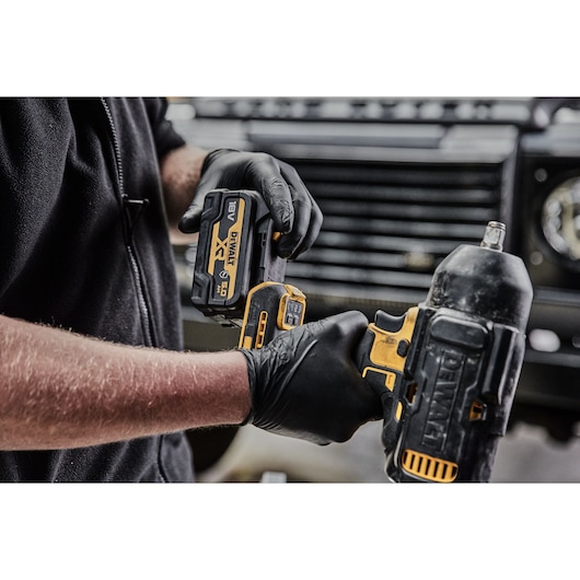 A person wearing black gloves is attaching a DEWALT 18V XR battery to a DEWALT cordless power tool, with a vehicle in the