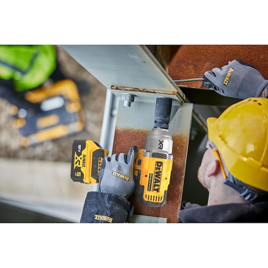 A close-up view of a construction worker using a DEWALT cordless impact wrench on a steel beam, wearing safety gloves and a yellow hard hat. Another worker is blurred in the background holding a DEWALT tool.