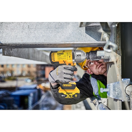 A person wearing safety gloves and a helmet is using a DEWALT DCF900P2T cordless impact wrench to fasten a large bolt on a metal structure at an outdoor construction site.