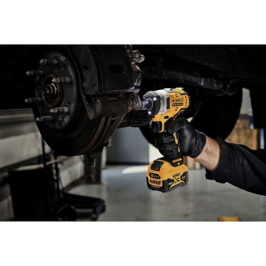 A person wearing a black glove uses a yellow DEWALT cordless impact wrench to work on a car's brake system in a garage