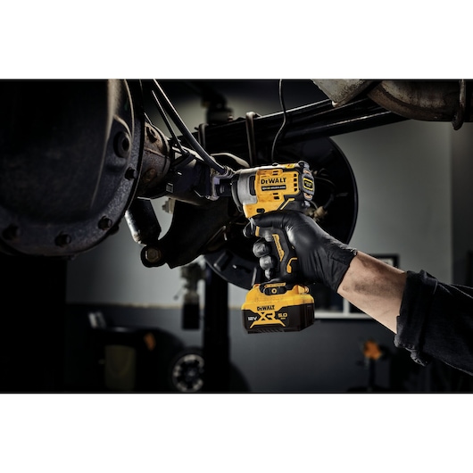 A person wearing a black glove uses a yellow DEWALT DCF901P1 12V XR cordless impact wrench to work on a vehicle's suspension