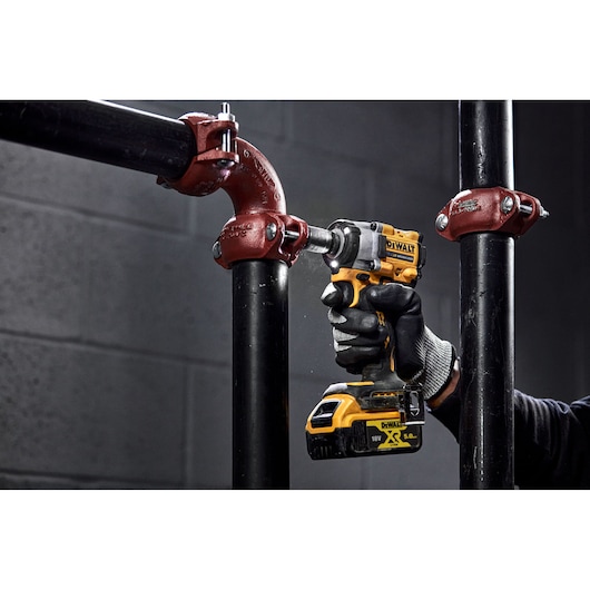 A person wearing gloves uses a DEWALT DCF921NT cordless impact wrench to tighten or loosen a metal pipe fitting in an industrial setting.
