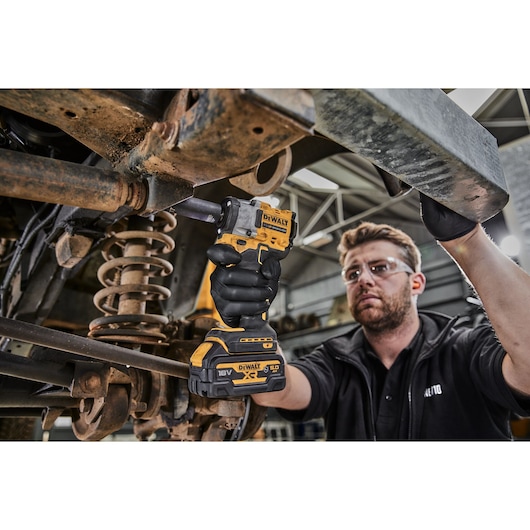 A person is using a DEWALT DCF921NT cordless impact wrench to work on the suspension of a vehicle in a workshop. The worker's face is blurred and they are wearing black gloves.