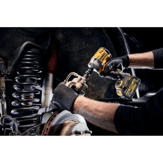 A person wearing black gloves uses a DEWALT cordless impact wrench to work on a vehicle's suspension and brake components.