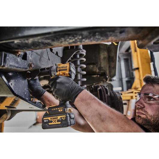 A person wearing black gloves using a DEWALT 18V XR cordless impact wrench to work on the underside of a vehicle in an automotive workshop.
