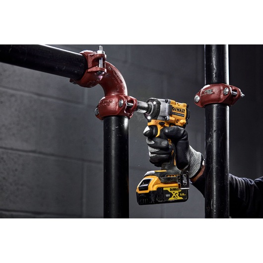 A person wearing black gloves uses a yellow and black DEWALT cordless impact wrench to tighten a bolt on a metal pipe fitting in an industrial setting.