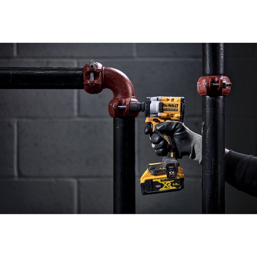 A person wearing a black and grey work glove is using a DEWALT 18V cordless impact wrench on a black metal pipe fitting in an industrial setting. The background is a grey brick wall.