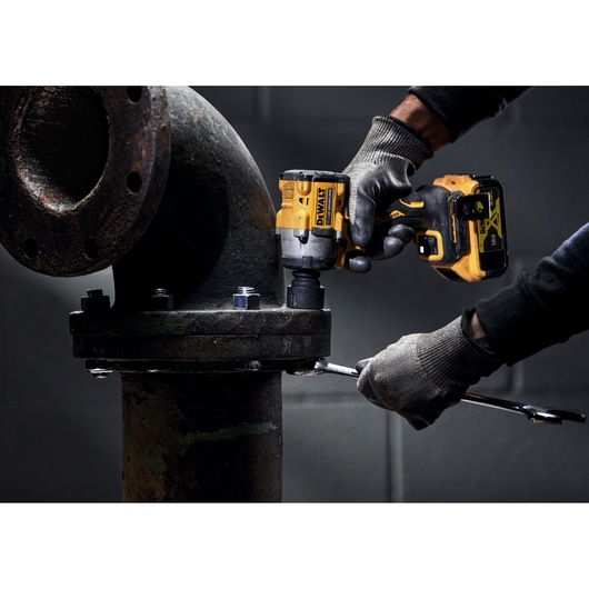 A person wearing gloves is using a DEWALT cordless impact wrench to tighten bolts on a large metal industrial pipe fitting.